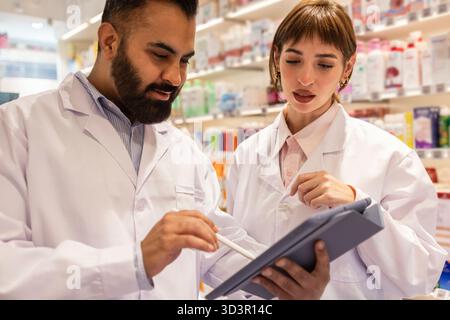 Zwei Apotheker in Labormänteln konsultieren ein digitales Tablet in einer Apotheke und besprechen Rezepte und Patientenversorgung. Regale gefüllt mit Medikamenten und Profis Stockfoto