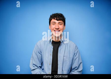 Ein junger Mann mit Zahnspangen lächelt warm in die Kamera, trägt ein Jeansshirt über einem schwarzen T-Shirt. Der glatte blaue Hintergrund unterstreicht sein fröhliches DE Stockfoto