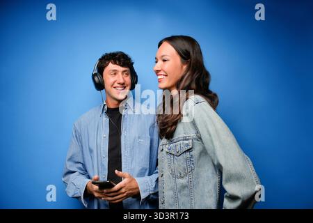 Ein junger Mann und eine junge Frau genießen gemeinsam Musik, lächeln und plaudern. Der Mann trägt Kopfhörer und hält ein Smartphone vor einem hellblauen Hintergrund Stockfoto