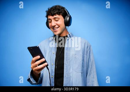 Ein junger Mann mit Kopfhörern lächelt, während er Musik auf seinem Handy vor einem leuchtend blauen Hintergrund genießt. Seine lässige Kleidung und sein entspanntes Verhalten reflektieren Stockfoto