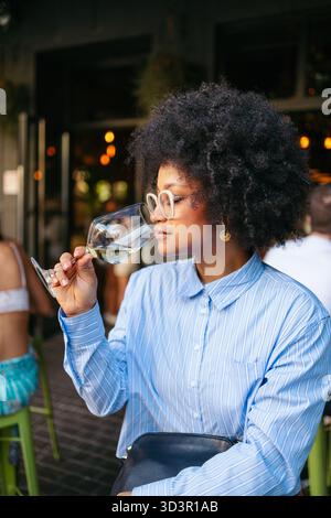 Eine Frau mit stilvollen Gläsern genießt ein Glas Weißwein in einem gemütlichen Straßencafé in einer belebten Innenstadt, umgeben von urbanem Charme und Abend A Stockfoto