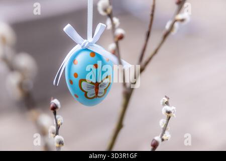 Ein leuchtend blaues Osterei, das mit einem Schmetterling und Polka Dots geschmückt ist, hängt an den blühenden Zweigen. Das rustikale Dekor wird durch das zarte weiße Band verstärkt Stockfoto