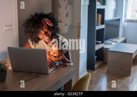 Afroamerikanische Mutter und Tochter verbrachten zu Hause gute Zeit zusammen, mit einem Laptop auf einem Holztisch und schaffen eine warme und liebevolle fami Stockfoto