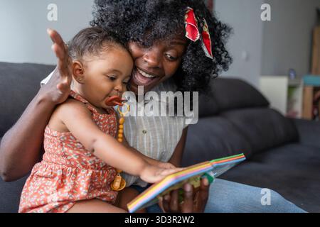 Afroamerikanische Mutter und Tochter genießen eine gute Zeit zusammen auf der Couch zu Hause. Die Mutter liest ihrer Tochter vor und schafft ein warmes und n Stockfoto