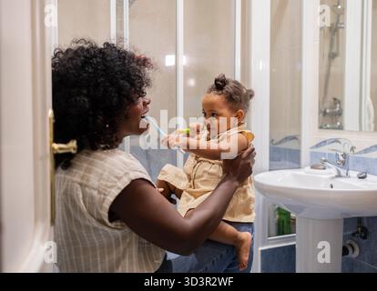 Afroamerikanische Mutter und Tochter teilen sich einen freudigen Moment zu Hause, während sie sich im Badezimmer die Zähne putzen, die Zahnhygiene fördern und Stockfoto