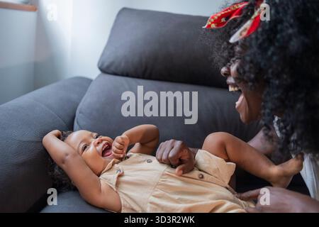 Afroamerikanische Mutter und Tochter lachen zu Hause zusammen und halten einen fröhlichen und liebevollen Moment auf einem bequemen Sofa fest. Herzliche Familienbindung und Stockfoto