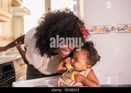 Afroamerikaner Mutter und Tochter genießen eine gute Zeit zu Hause in der Küche. Die Mutter lacht, während ihre Tochter glücklich auf einem leckeren Te isst Stockfoto