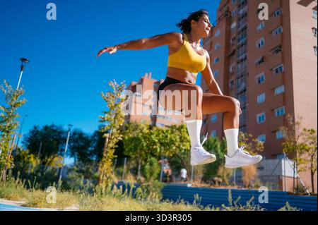 Der niedrige Winkel einer Sportlerin in Sportausrüstung führt während des Outdoor-Trainings an einem hellen Tag einen explosiven Sprung durch. Der städtische Hintergrund ist grün Stockfoto