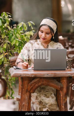 Eine muslimische Frau in einem wunderschönen Kleid mit Öko-Print arbeitet an ihrem Laptop an einem rustikalen Holztisch. Die Naturfärbekleidung unterstreicht nachhaltige Mode und Stockfoto