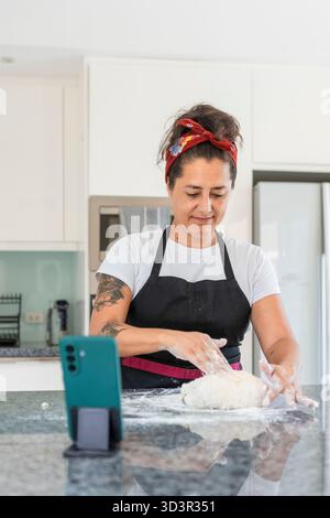 Frau knete Teig auf einer Arbeitsplatte, bastelte köstliches hausgemachtes Brot in einer modernen Küche. Ein Smartphone befindet sich in der Nähe, möglicherweise für ein Rezept oder r Stockfoto