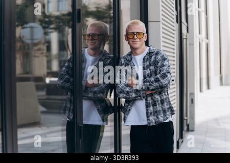 Ein junger Mann mit Albino-Aussehen posiert selbstbewusst vor einem Glasfenster und spiegelt urbane Architektur wider. Er trägt eine stilvolle Sonnenbrille und einen Casua Stockfoto