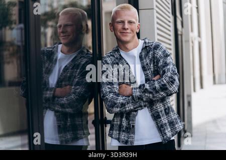 Ein junger Mann mit Albino-Aussehen steht selbstbewusst vor einem modernen Gebäude, seine Arme überkreuzt und ein warmes Lächeln im Gesicht, das eine positive Haltung widerspiegelt Stockfoto