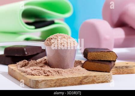 Eine Nahaufnahme von mit Schokolade überzogenen Proteinriegeln und eine Schaufel Proteinpulver auf einem Holzbrett. Die Trainingsgeräte im Hintergrund stellen eine Fitness dar Stockfoto