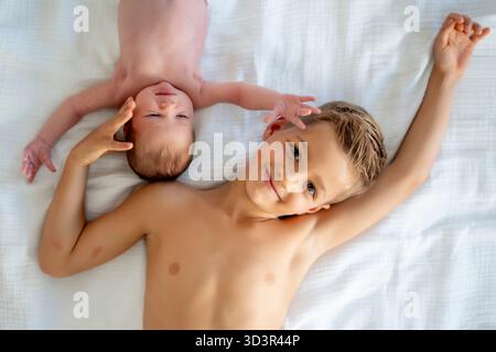 Zwei Geschwister liegen zusammen auf weißen Bettlaken und teilen einen freudigen Moment. Der ältere Bruder lächelt, während das Baby bequem ausruht, und hebt das hervor Stockfoto