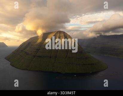 Eine atemberaubende Landschaft eines majestätischen Berges auf den Färöern, getaucht in warmes Sonnenuntergangslicht. Die majestätischen Gipfel erheben sich inmitten eines ruhigen Meeres, mit Drama Stockfoto