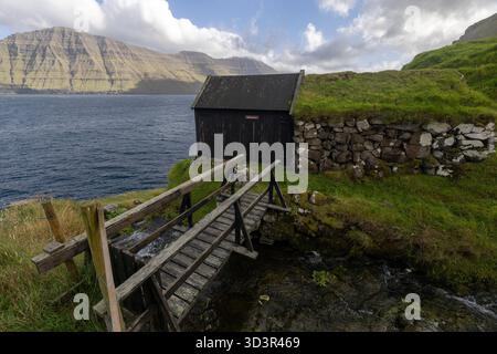 Eine malerische Szene der Färöer-Inseln zeigt eine rustikale Hütte mit einem Grasdach in der Nähe des Ozeans. Eine Holzbrücke überquert einen kleinen Bach, mit Stockfoto