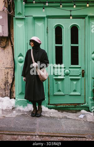 Frau im Mantel und rosa Hut lächelt mit grüner Tür in städtischer Umgebung Stockfoto