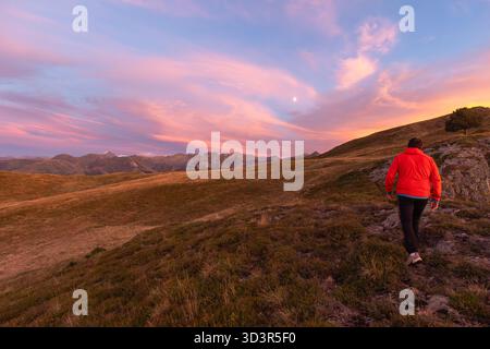 Ein Mann in einer roten Jacke spaziert auf einem grasbewachsenen Hügel während eines atemberaubenden Sonnenuntergangs in den Pyrenäen. Der Himmel ist mit rosa und orangen Tönen bemalt, was eine Ruhe schafft Stockfoto