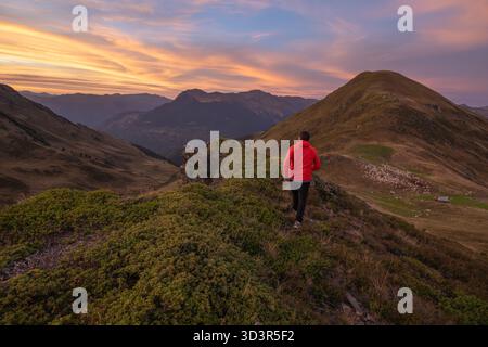 Ein Mann in roter Jacke spaziert während eines atemberaubenden Sonnenuntergangs in den Pyrenäen auf einem Bergpfad. Der lebendige Himmel und die sanften Hügel schaffen ein atemberaubendes n Stockfoto