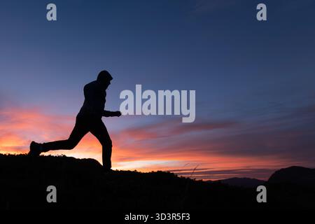 Ein Mann in Silhouette wird vor der atemberaubenden Kulisse eines pulsierenden Sonnenuntergangs in den Pyrenäen gefangen. Der Himmel ist mit Orangentönen bemalt, Stockfoto