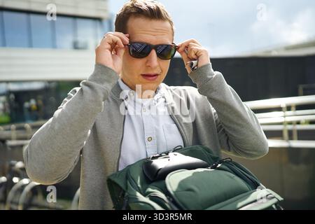 Ein Mann, der eine Sonnenbrille trägt, passt sie an, während er draußen steht. Er trägt legere Kleidung mit einem grauen Pullover und trägt einen grünen Rucksack unter einem cl Stockfoto