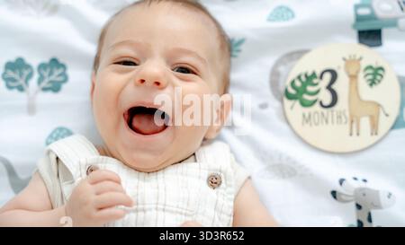 Ein süßes 3 Monate altes Baby in leichter Kleidung, liegt auf einer gemütlichen bedruckten Decke und lächelt fröhlich. Die hellen Augen und das Lächeln des Babys fangen einen Moment des pur ein Stockfoto