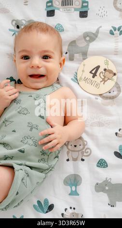 Ein lächelndes, vier Monate altes Baby liegt auf einer verspielten Decke im Safaristil und markiert mit einem hölzernen Tafelaufkleber einen freudigen Meilenstein in ihrer Entwicklung Stockfoto