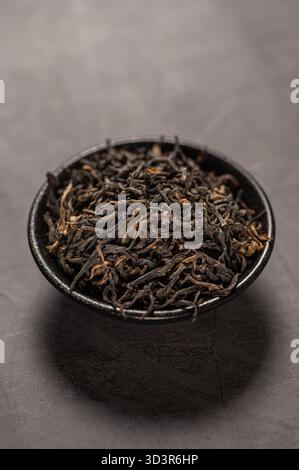 Ein Nahbild von Dianhong trockenen Teeblättern, die elegant in einer schwarzen Keramikschale platziert sind. Die kräftigen Braun- und Schwarztöne unterstreichen die erstklassige Qualität des Tees Stockfoto