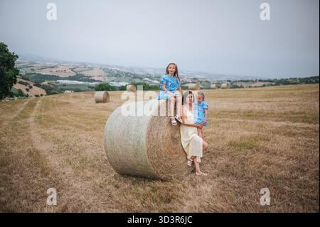 In einem ruhigen Moment auf dem italienischen Land sitzen eine Mutter und ihre Töchter auf einem Heuballen. Sanfte Hügel und klarer Himmel schaffen ein pict Stockfoto