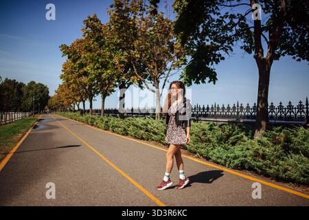 Eine junge Frau läuft einen sonnigen Parkweg entlang, umgeben von üppigem Grün und Bäumen. Sie ist lässig gekleidet, genießt die friedliche Atmosphäre und einen Sen Stockfoto
