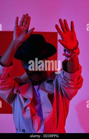 Porträt eines afroamerikanischen Mannes mit Schnurrbart in modischem Trenchcoat, Fedorahut und Sonnenbrille, die in Neonlicht tanzt Stockfoto