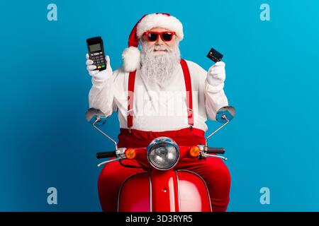 Der Weihnachtsmann auf einem roten Motorrad hält eine Kreditkarte und ein Smartphone als verspielte Festszene für Weihnachten und Weihnachtseinkäufe Stockfoto