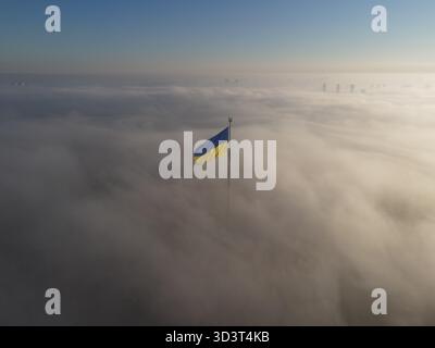 Ukrainische Nationalflagge, die an einem nebeligen Morgen in Kiew, Ukraine, über den Wolken fliegt. Drohnenansicht aus der Luft. Stockfoto