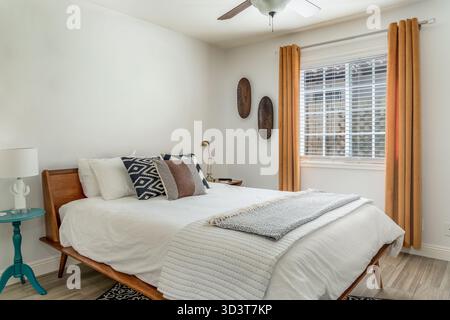 Modernes Wüstenschlafzimmer mit Holzrahmenbett, Akzentkissen, Decken, Lampe und Fenster mit Vorhängen Stockfoto