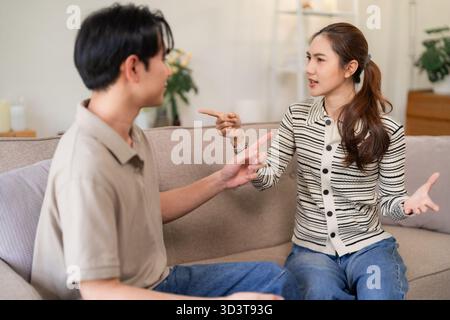 Asiatisches erwachsenes Paar, das zu Hause intensive häusliche Auseinandersetzungen erlebt. Partner äußern Frustration, Meinungsverschiedenheiten und Spannungen während eines ernsthaften Gesprächs auf dem Sofa. Stockfoto