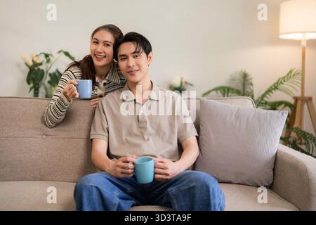 Fröhliche asiatische Paare teilen warme Getränke auf einem komfortablen Sofa zu Hause und entspannen gemeinsam in einem gemütlichen hellen Wohnbereich Stockfoto