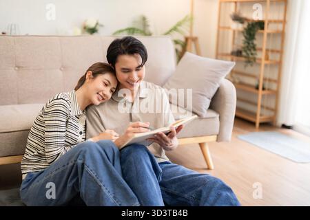 Asiatisches Erwachsenenpaar genießt eine komfortable häusliche Umgebung. Der Mann schreibt im Buch, die Frau ruht den Kopf. Angenehme, friedliche Alltagsszene. Fröhlich harmonisch Stockfoto