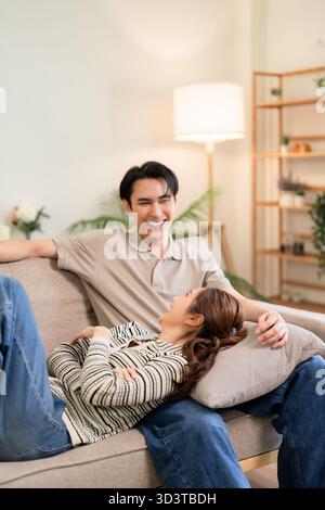 Glückliches asiatisches Paar genießt Entspannung zu Hause. Sie teilen sich lächelnden Komfort und eine liebevolle Verbindung auf einem gemütlichen Sofa in ihrem Wohnbereich. Stockfoto