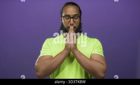Mann trägt ein neongrünes Hemd mit zusammengedrückten Händen im Gebet in einem lila Studio-Setting; Achtsamkeit ruhig. Stockfoto