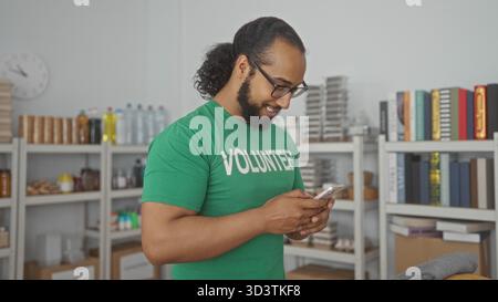 Ein Freiwilliger hält ein Smartphone und tippt mit beiden Händen im Spendengebäude, lächelt beim Lesen von Nachrichten; Unterstützung der Mitgefühl-Community. Stockfoto