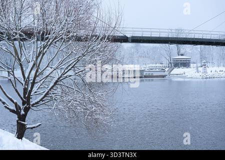 Im Schnee, eine Brücke über den Fluss am Straßenrand, eine Kabelbrücke, große Schneeflocken, Schneehaufen am Straßenrand, ein PA Stockfoto