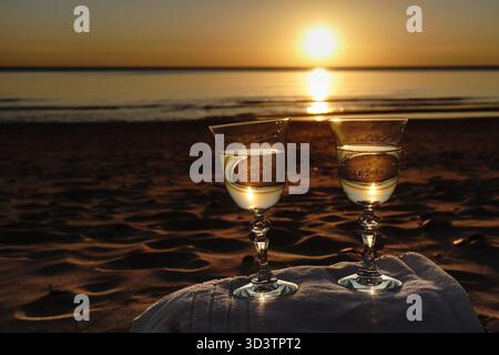 Zwei Gläser Wein am Meer bei Sonnenuntergang, Weißwein in Gläser gegossen, ein improvisierter Tisch mit einem Hotelhandtuch am Sandstrand, ein Ball der se Stockfoto