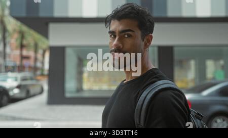 Mann in schwarzem Hemd mit Rucksack, der über die Schulter auf die Stadtstraße schaut, indem er eine Fassade inmitten verschwommener Autos baut; Neugier. Stockfoto