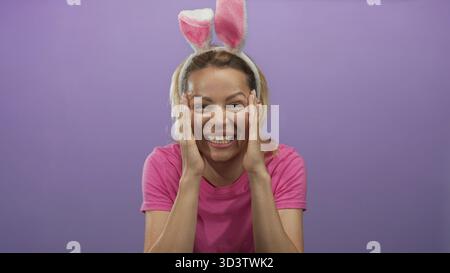 Frau hält die Wangen mit beiden Händen in einem lila Studio-Setting mit Hasenohren und rosa Hemd; Überraschung. Stockfoto