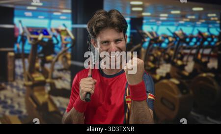 Mann, der Tennisschläger über die Schulter packt und geballte Faust in der Turnhalle mit Laufbändern und Heimtrainer in der Nähe pumpt; Siegesbestimmung. Stockfoto