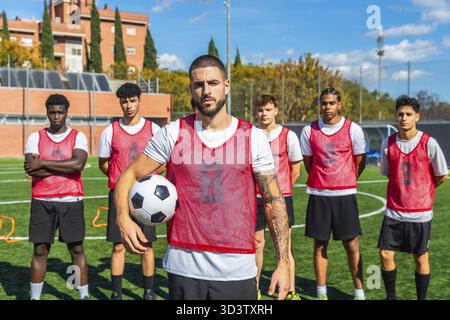Fußballkapitän steht mit Ball auf grünem Feld und führt ein vielfältiges, entschlossenes Team junger männlicher Spieler in Training und Vorbereitung auf den Wettkampf o Stockfoto