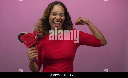 Frau mit Klebebandspender und flexiblem Bizeps im Studio mit rosa Hintergrund und breitem lachenden Lächeln; Selbstvertrauen-Feier. Stockfoto