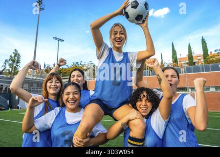 Eine Gruppe verschiedener Fußballspielerinnen jubeln und heben eine Teamkollegin mit einem Fußball auf einem grünen Feld an, um Glück und Teamarbeit nach einem C zum Ausdruck zu bringen Stockfoto