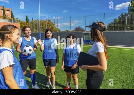 Fußballspielerinnen in blauen Pinnies hören aufmerksam zu, wie ihre Trainerin Strategien erklärt und die Mannschaft auf einem sonnigen Kunstgras führt Stockfoto