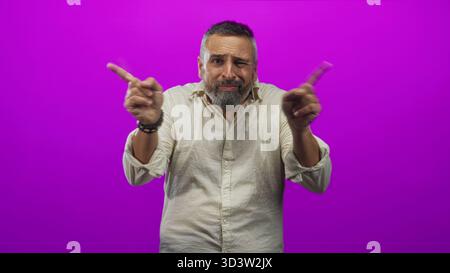 Mann mit grauem Bart, Sträucher und Spitzen mit offenen Handflächen in lila Studio, mit beigem Hemd und perlenbesetzten Armbändern; Ungewissheit. Stockfoto